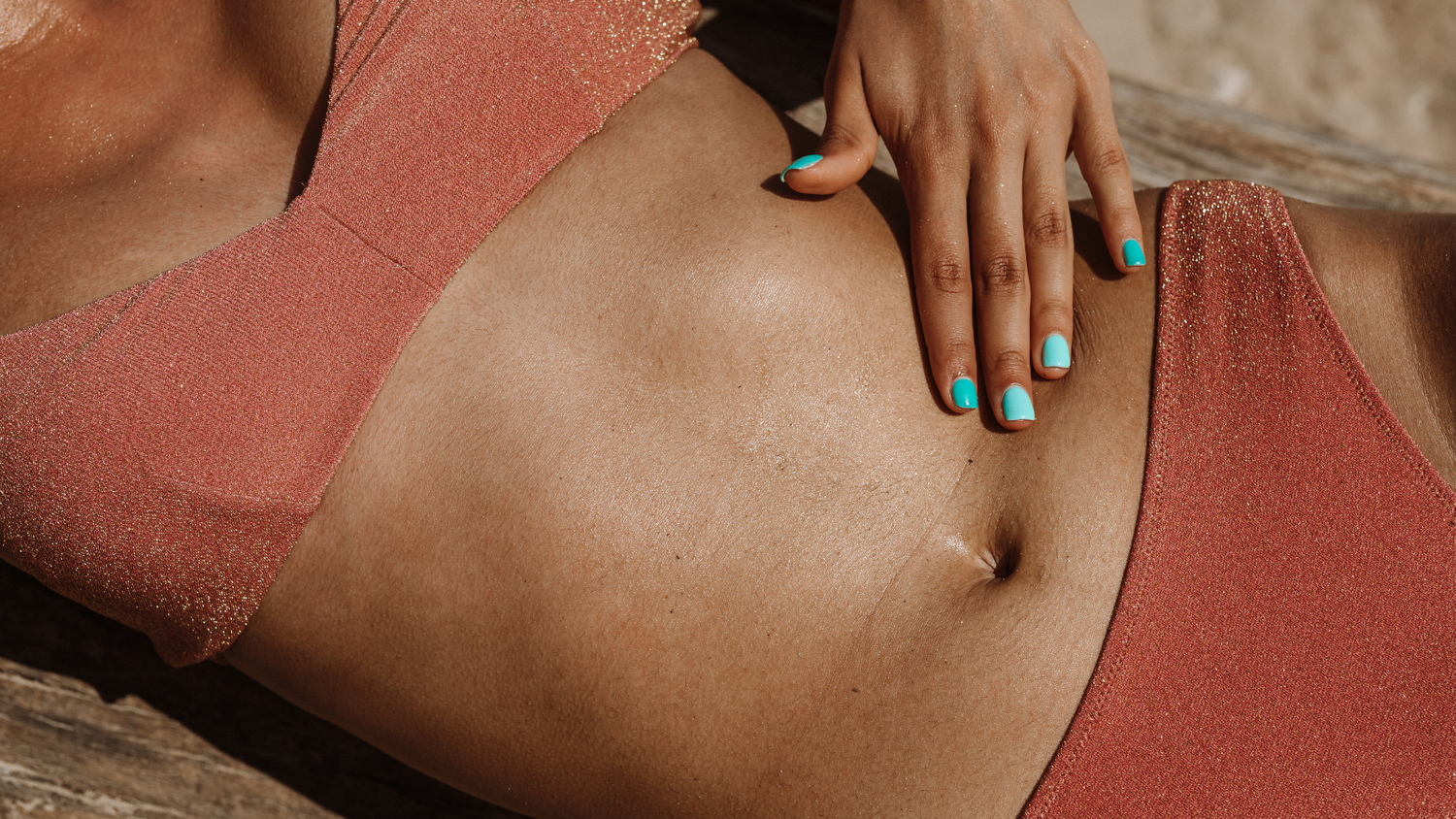 corps d'une femme bronzée avec un maillot de bain terracotta se touchant le vendre avec sa main gauche dont les ongles sont vernis bleu pastel