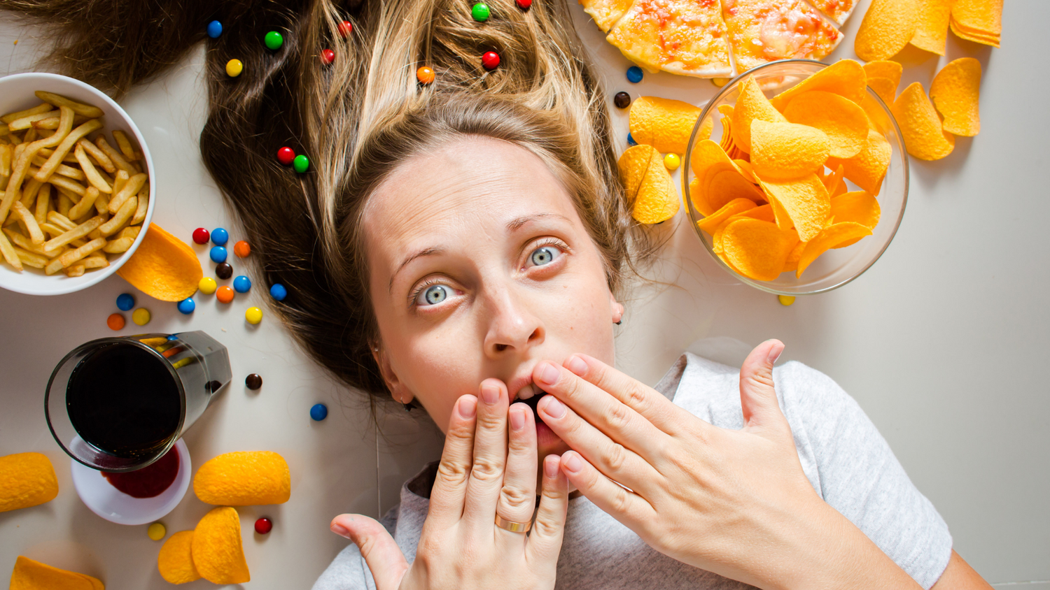 Femme blonde allongée au milieu de nourriture grasse et peu saine (chips, frites, pizza)les mains sur la bouche pour dire oups