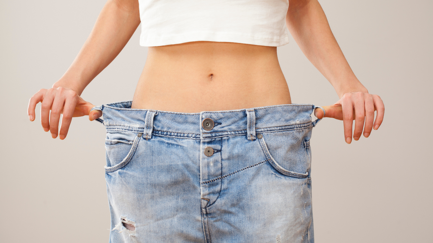 femme mince portant un croptop blanc et un jean qu'elle étire avec ses deux mains de chaque côté pour signaler une perte de poids