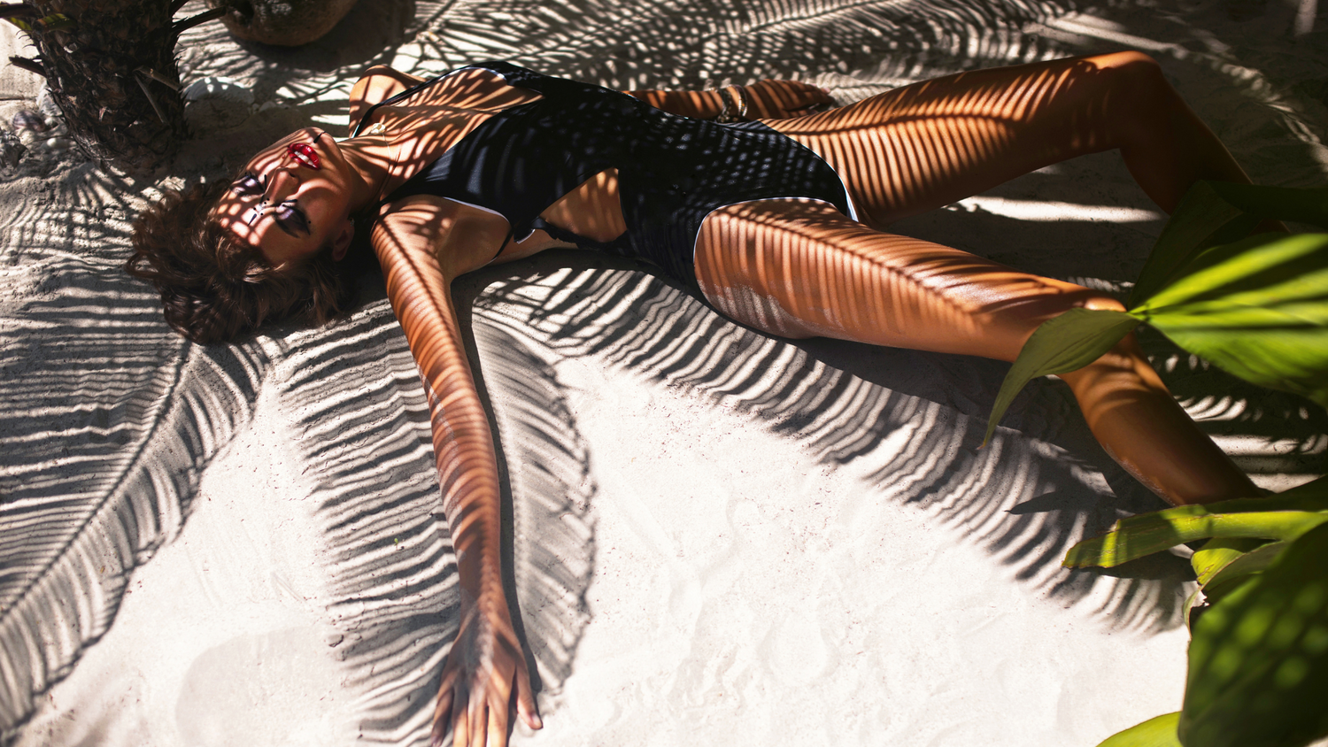 femme brune mince en maillot de bain une pièce noir allongée dans le sable bras écartée avec l'ombre des feuilles de palmier artistiquement dessinées sur son corps
