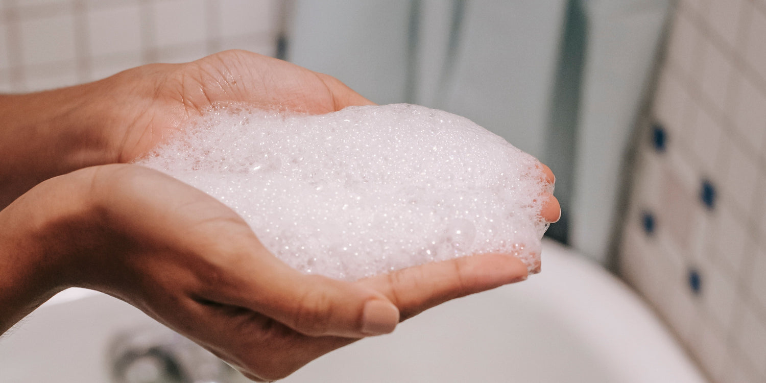 2 mains contenant de la mousse d'un shampoing chimique riche en tensioactifs au dessus d'une baignoire dans une salle de bain